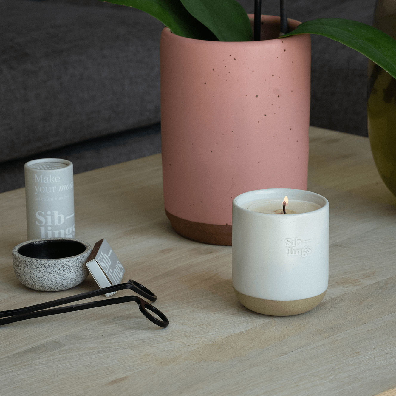 Lit Siblings candle in White Cloud vessel on light wooden table with match tube, small bowl, and black wick trimmers, soft pink plant pot in background