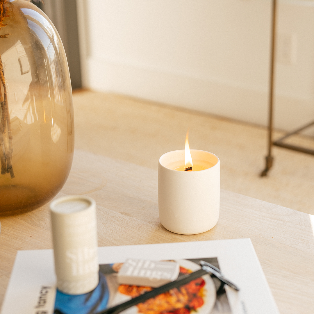 Lit No. 01 Siblings candle in a minimalist white vessel on a light wooden table, with a matchbox, trimmer, and amber vase nearby, creating a cozy, modern home scene.
