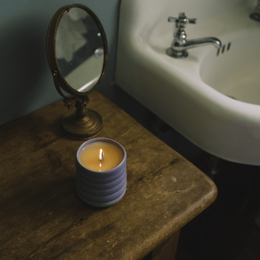 A lit candle in a Siblings wavy Forever Vessel sits on a rustic wooden surface next to a vintage brass-framed mirror.