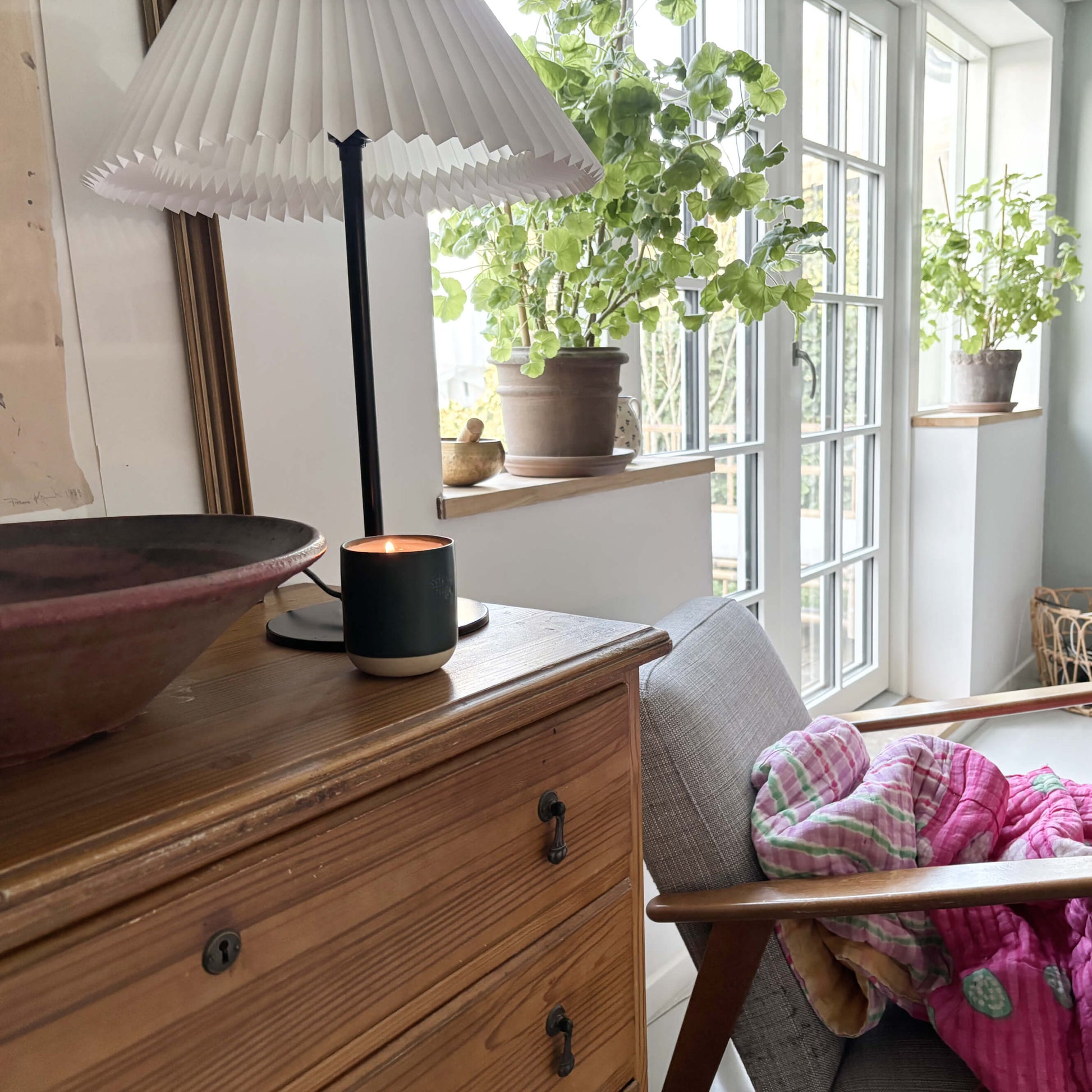 Lit Neroli Noir candle in a evergreen vessel on a rustic wooden dresser, next to a pleated white lamp and with green houseplants by large windows in the background, creating a cozy living space.