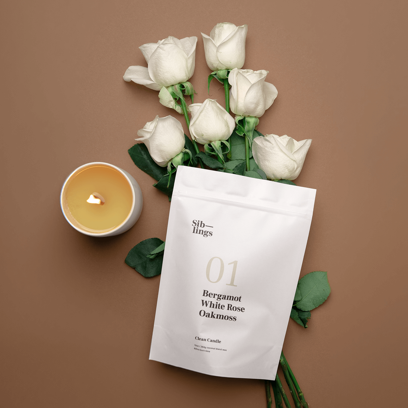 Lit No. 01 Siblings candle in a white vessel next to a white refill bag labeled "01 Bergamot White Rose Oakmoss" and a bouquet of white roses.