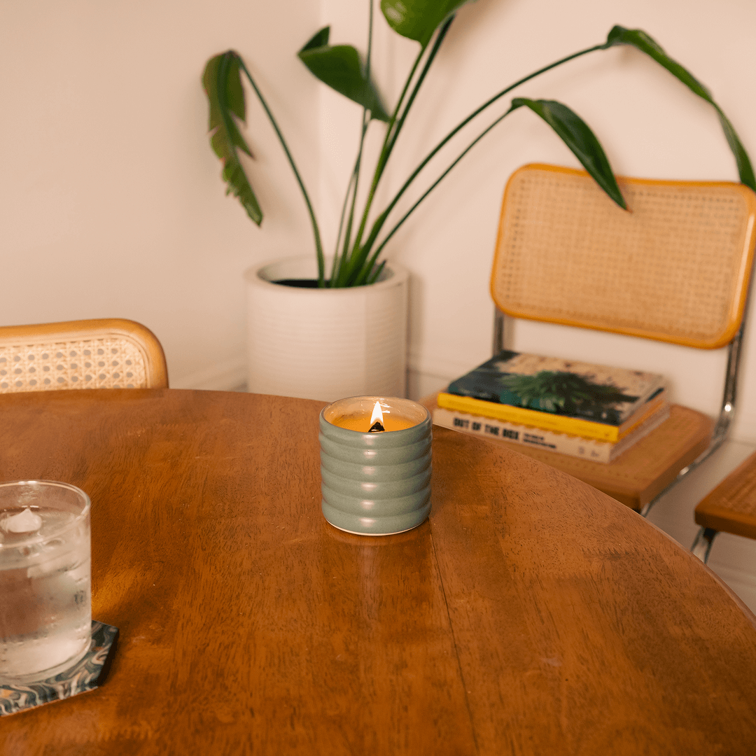 Lit No. 04 Siblings candle in a wavy green ceramic vessel on a wooden table, with a houseplant and a modern chair in the background, creating a cozy, natural home scene.