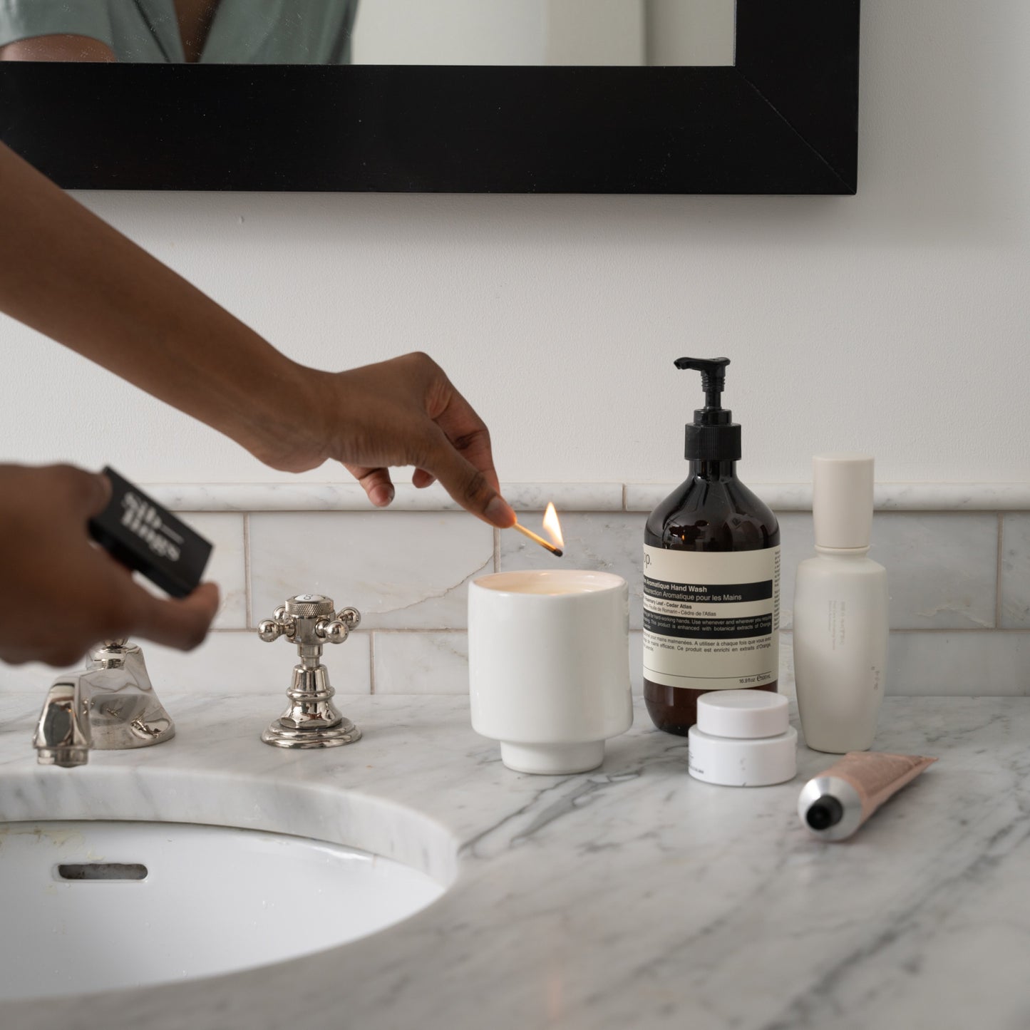 Person lighting a white Siblings candle in a modern ceramic holder on a marble bathroom countertop, and beauty products nearby, creating a cozy self-care scene.