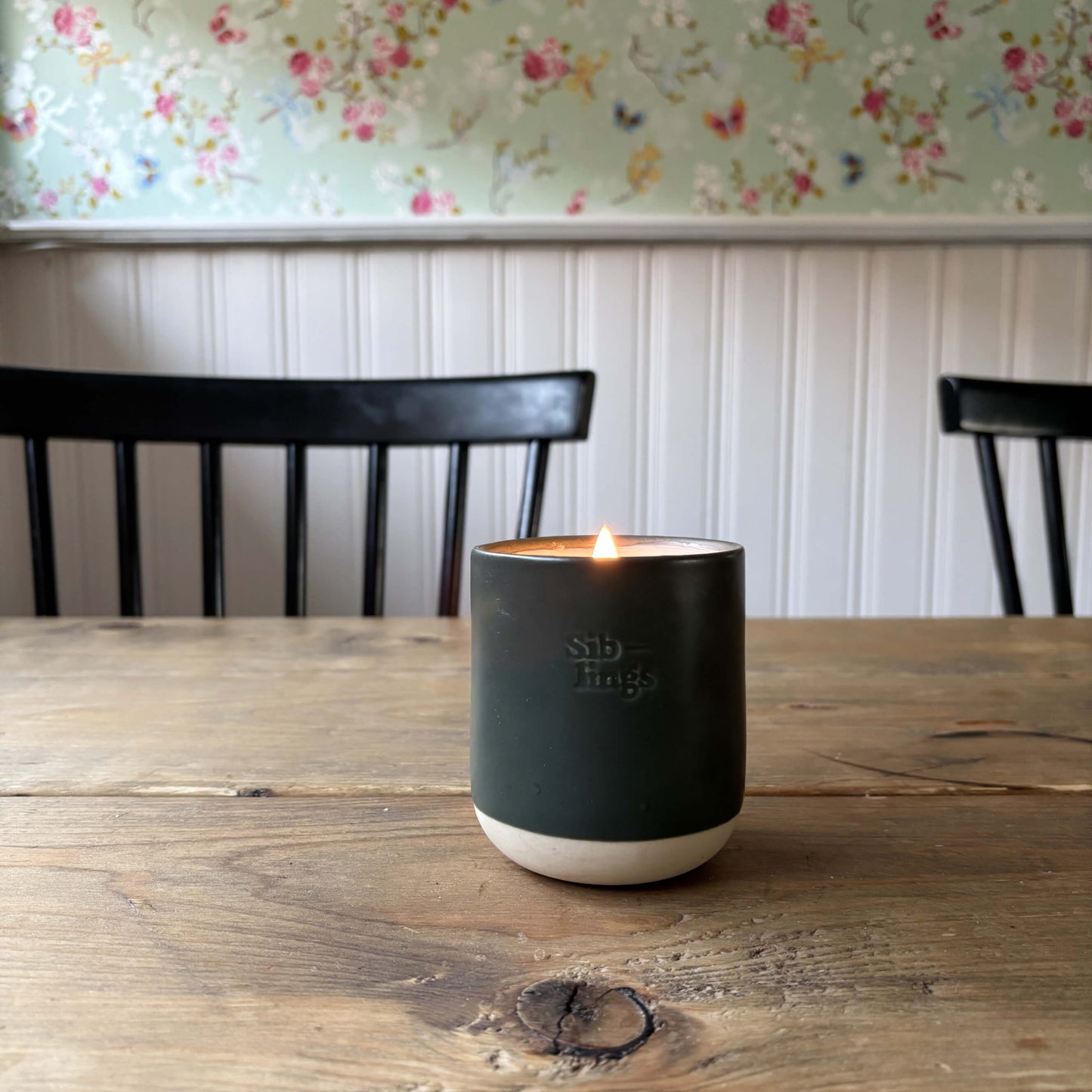 Lit Siblings candle in evergreen vessel on rustic wooden table, soft glow highlighting floral wallpaper and black chairs in the background. 