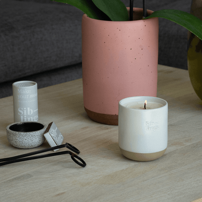 Lit Siblings candle in White Cloud vessel on light wooden table with match tube, small bowl, and black wick trimmers, soft pink plant pot in background