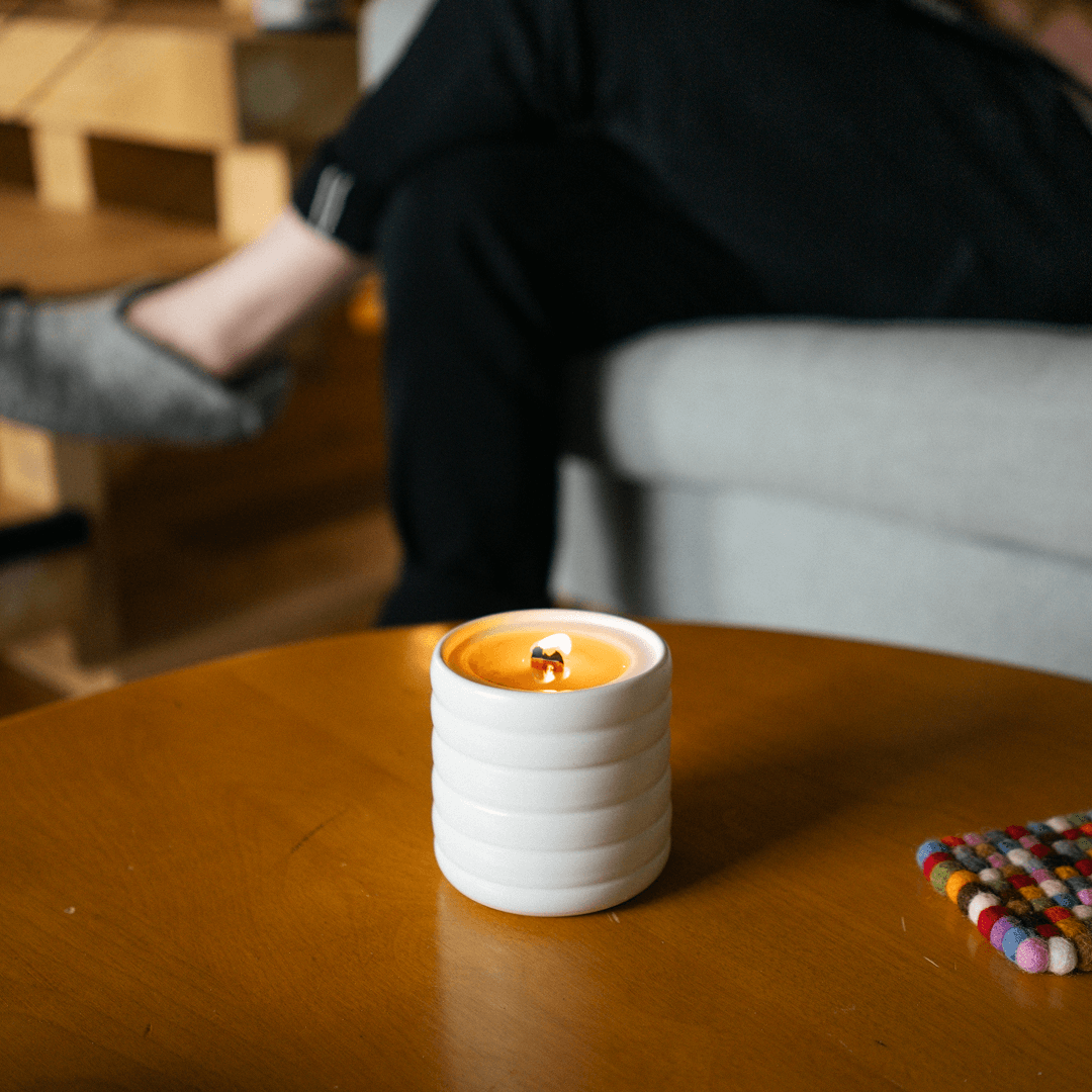 A lit candle in a sibling's white, wavy ceramic vessel sits on a wooden coffee table in a living room.
