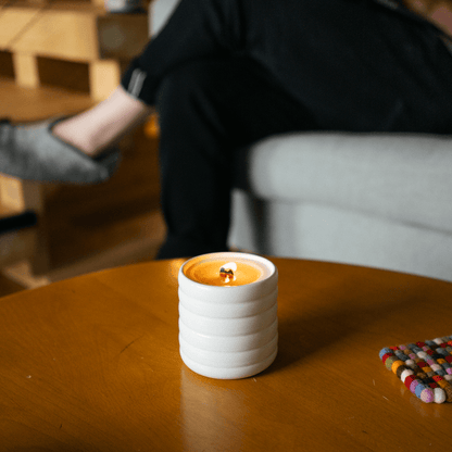 A lit candle in a sibling's white, wavy ceramic vessel sits on a wooden coffee table in a living room.
