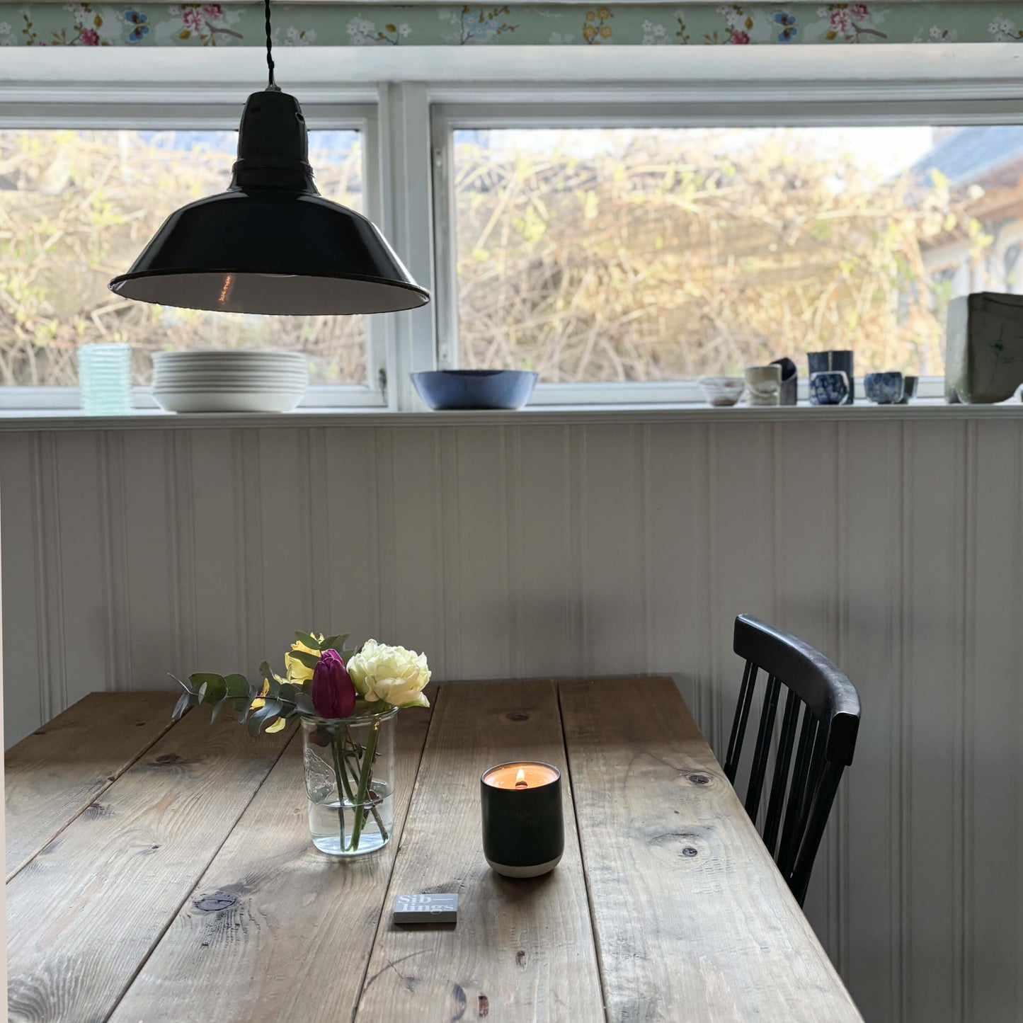 Rustic wooden dining table with yellow and pink flowers in a vase, a lit Siblings candle in a dark green siblings forever vessel, and a black industrial pendant lamp overhead, creating a cozy home ambiance.