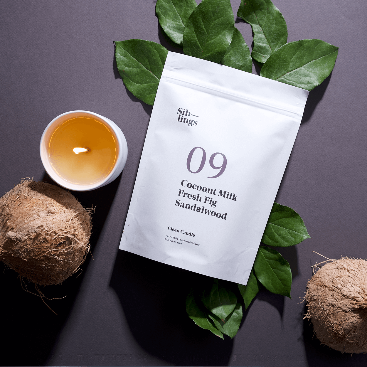 Lifestyle flat-lay of No. 09 — Coconut Milk, Fresh Fig, Sandalwood Siblings candle refill bag next to a lit candle, green leaves, and two whole coconuts, evoking a fresh, fruity, and cozy vibe.