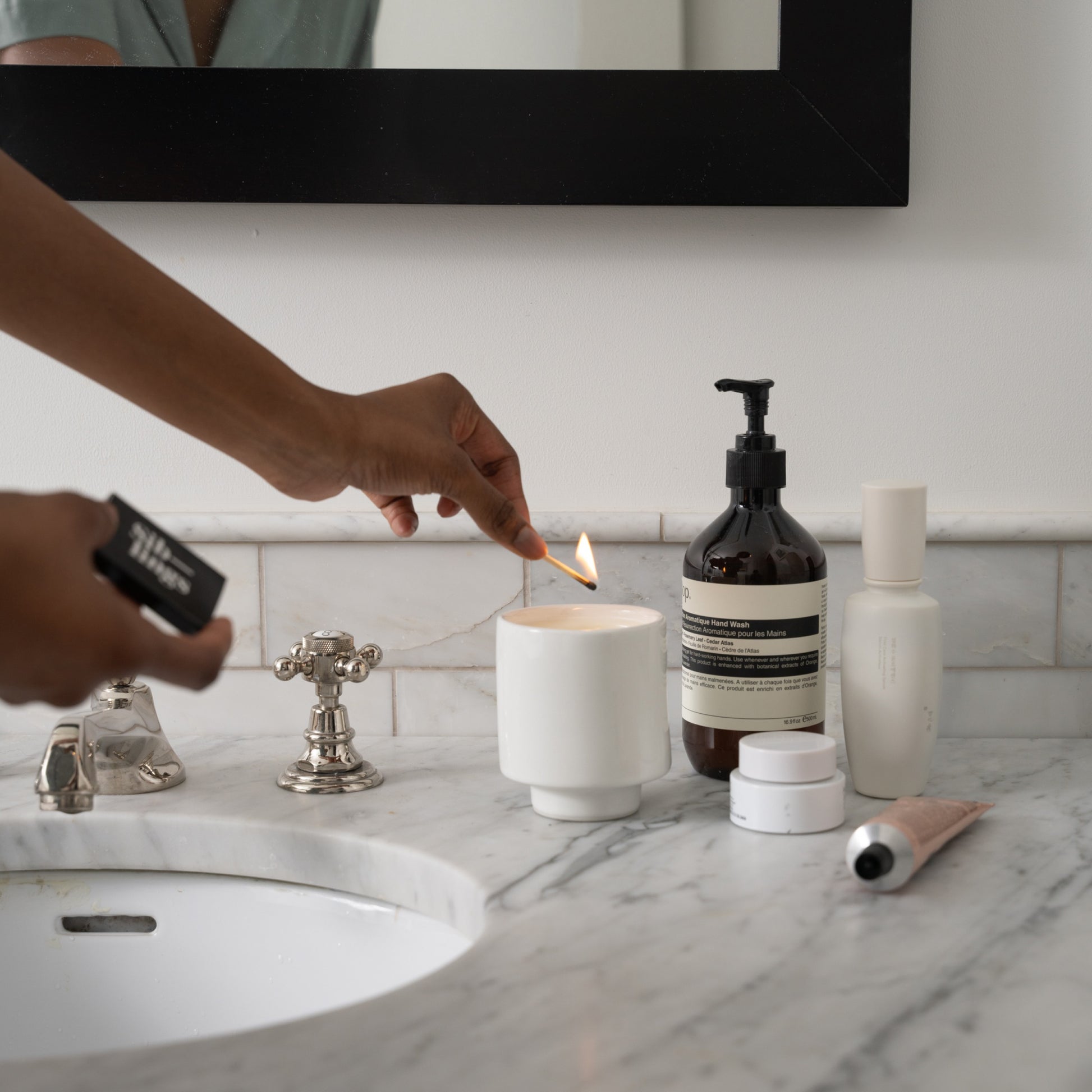 Person lighting a white Siblings candle in a modern ceramic holder on a marble bathroom countertop, and beauty products nearby, creating a cozy self-care scene.
