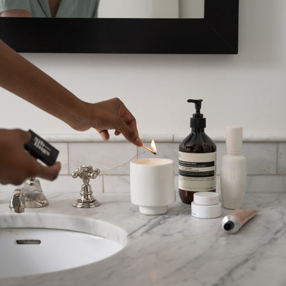 Person lighting a white Siblings candle in a modern ceramic holder on a marble bathroom countertop, and beauty products nearby, creating a cozy self-care scene.